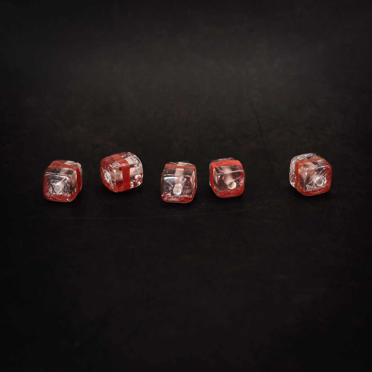 Fem rosa kubiska glaspärlor med röda rander uppradade, produktfotografi mot svart bakgrund