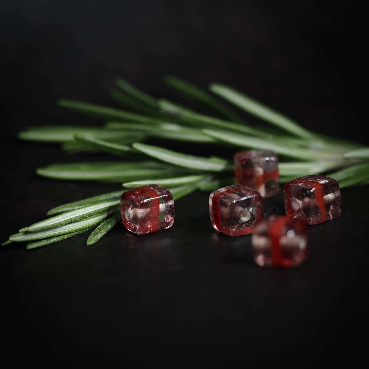 Rosa kubisk glaspärla med röd rand, fotograferad mot mörk bakgrund med barrkvister