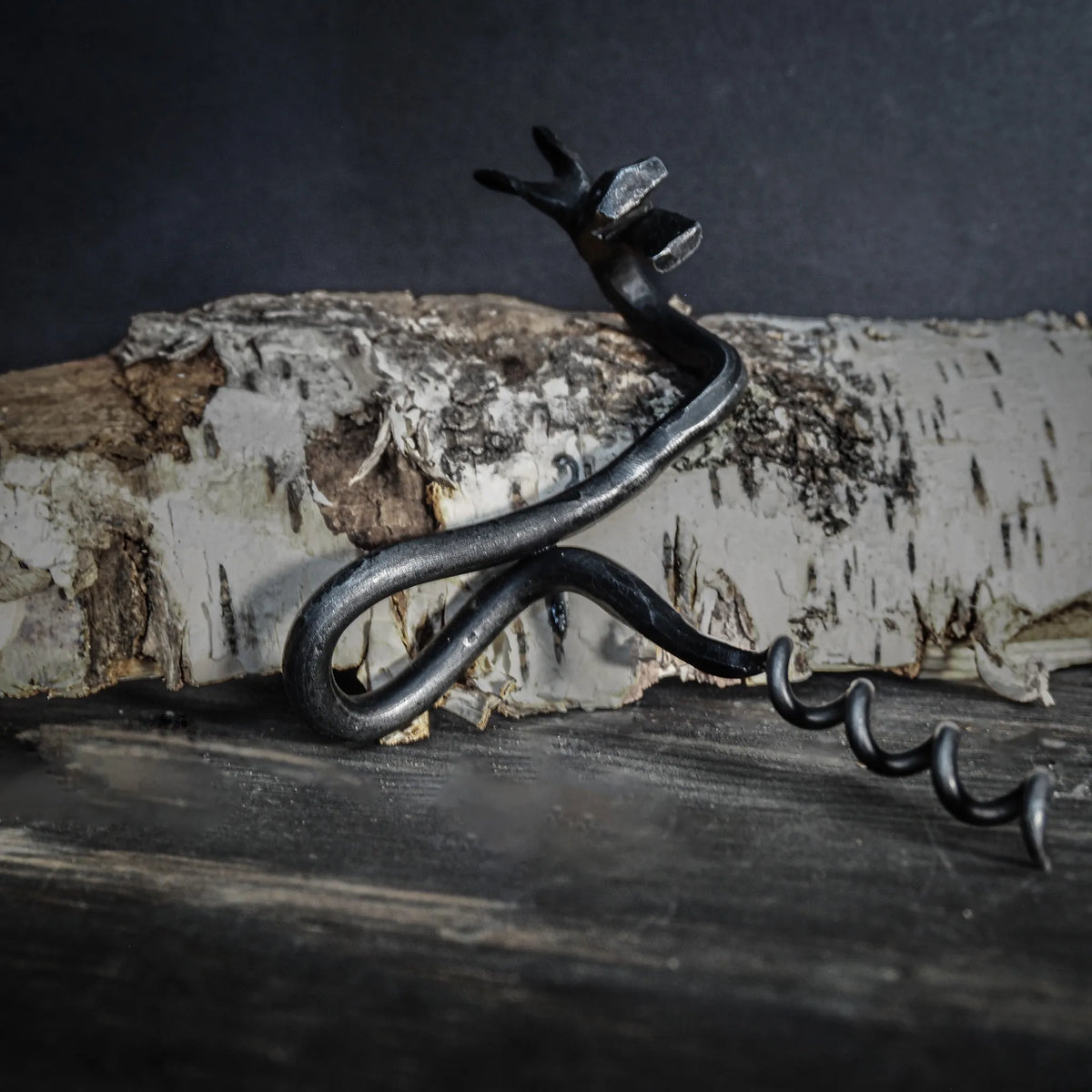 Detaljfoto av handsmide älgskruv. Älghuvudet visar realistiska ögon och detaljer i antikfinish. Metallhandtaget kurvar från älgskulpturen. Fotograferad mot mörkgrå bakgrund med dramatisk belysning som betonar smidets detaljer och textur.