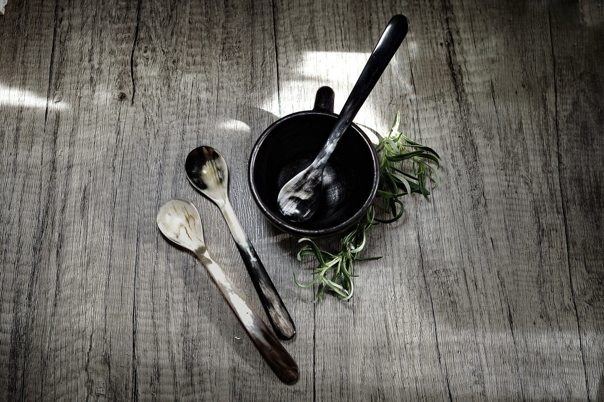 Lång tesked i flerfärgad ko-horn med naturliga mörkbruna och kremfärgade ränder, presenterad i en svart skål med färska rostmarv på grå trägolv.