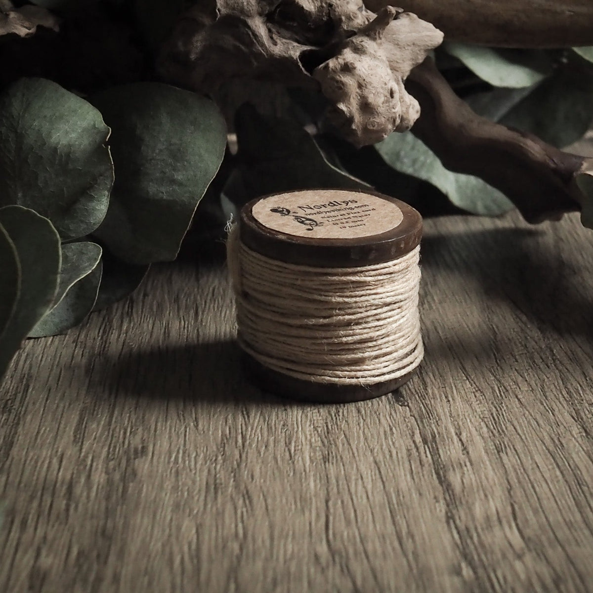 Linnetråd 0,5 mm på träspole med märkad etikett. Naturligt ljusgult linnetrådsmaterial omkring träbasen, presenterat på rustikt trägolv med torkade växter och grön lövverk i bakgrunden.