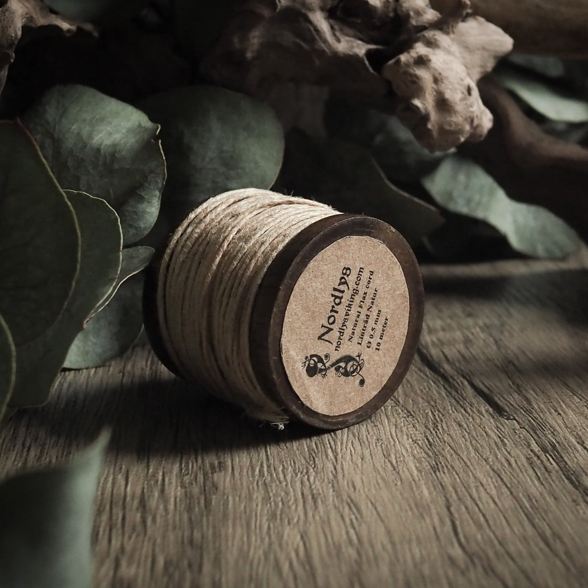 Linnetråd 0,5 mm på träspole med tydlig 'Nordlys' märkning på träetikett. Det kremfärgade linnetrådet är tätt lindad omkring träbasen. Presenterad med torkade grenar och grön eukalyptus för artisanalkänsla.