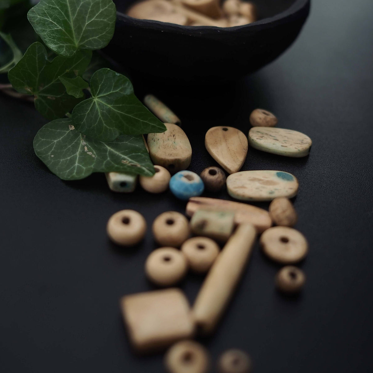 Blandning av vita benpärlor arrangerade på mörk yta med gröna växter. Pärlorna visar naturlig benåldrande och varierar mellan sfärer, oval formade och cylindriska rör med hand-snidtad asymmetrisk karaktär.