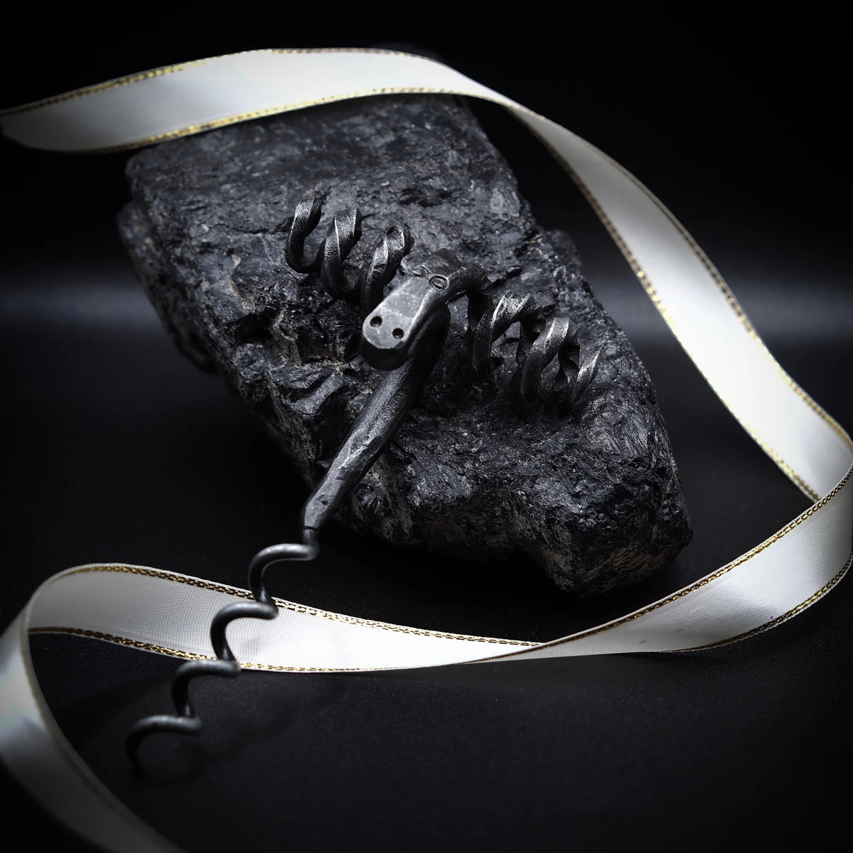 Bagge-skruv presenterad med silver/grå band med guldkant för lyx-packningseffekt. Ram-huvud visar detaljerat metallhantverk med synliga öga och horndetaljer. Dunkelstensunderlag understryker artisan-estetik. Kontrollerad belysning betonar både verktygets metallhantverk och presentationskvalitet. Tjänar både produktdokumentation och lyx-gåva-marknadsföring.