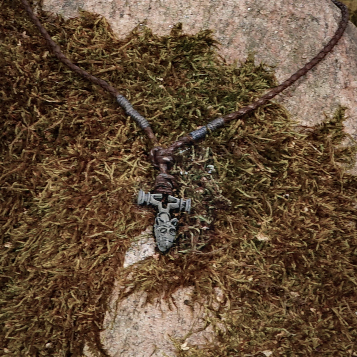 Vargkorset halsband fotograferat på naturlig stenunderlag med mossa. Silverfärgat tenn-kors med brunt läderband, visar den geometriska Viking-designen i naturligt ljus mot jordnära bakgrund.