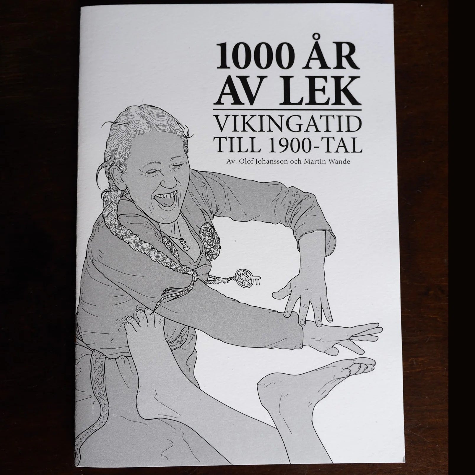 Häfte om '1000 År av Lek' från vikingatid till 1900-tal, med illustrerad omslag av Martin Wande, på mörk träbakgrund