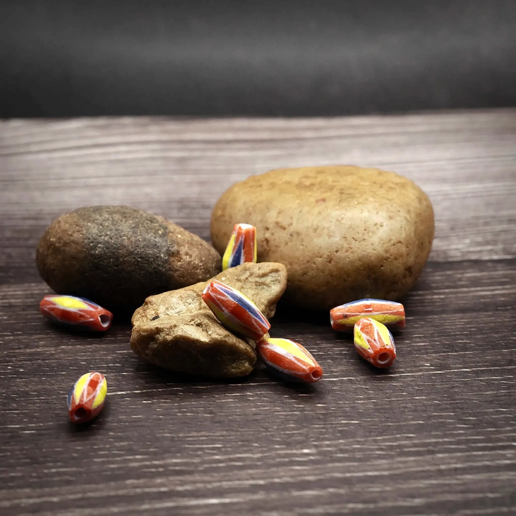 Multifärgad avlång glaspärla i rött, gult och orange (11mm längd, 3mm bredd, 1mm hål) fotograferad på mörkträ med naturliga stenar för skalreferens