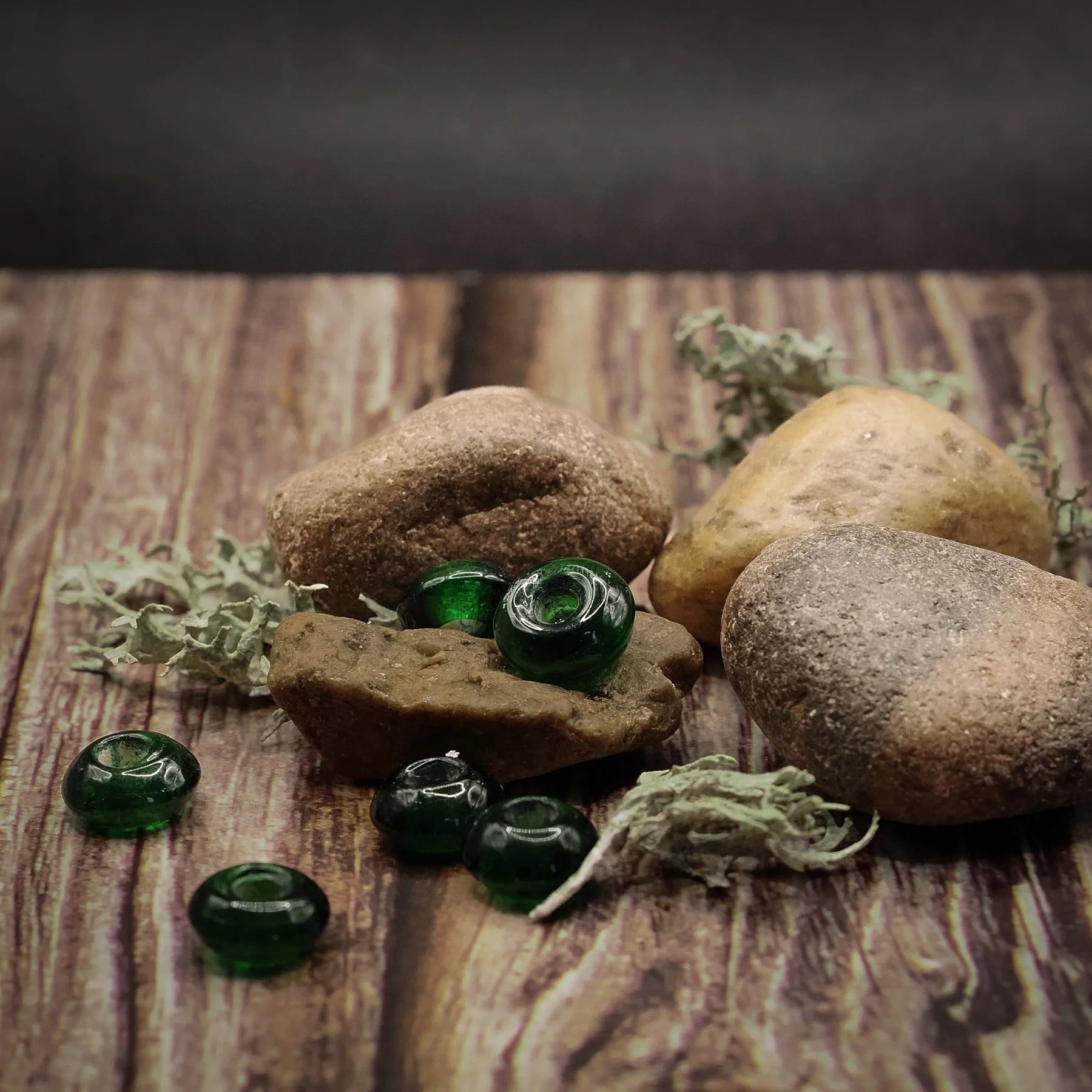 Gröna glaspärlor presenterade på rustikt trä med naturmaterial. Tre sfäriska pärlor visas tillsammans med ett träkärl och naturliga dekorativa element.