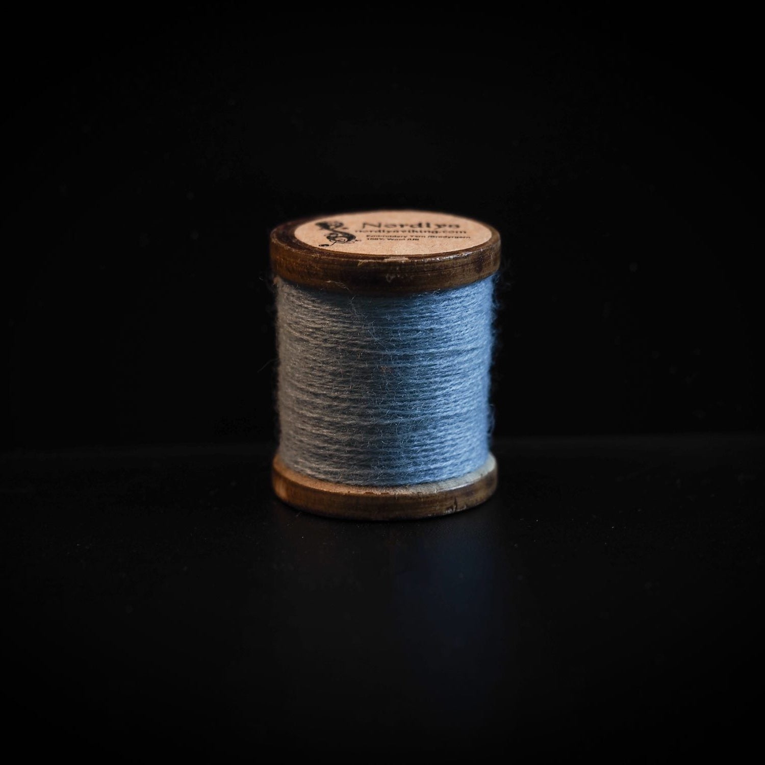 En enkel spole med blågrå ullgarn på mörk bakgrund, visar nordisk färgpalett och högkvalitativ brodyrtråd med träspole