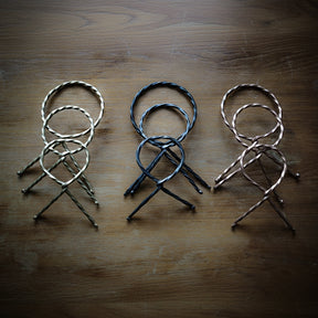 Tre hornställ i olika metallfinish (silver, svart, och koppar) arrangerade på rustik trävita bakgrund. Visar de vridna designen och metallets karakteristiska glans.