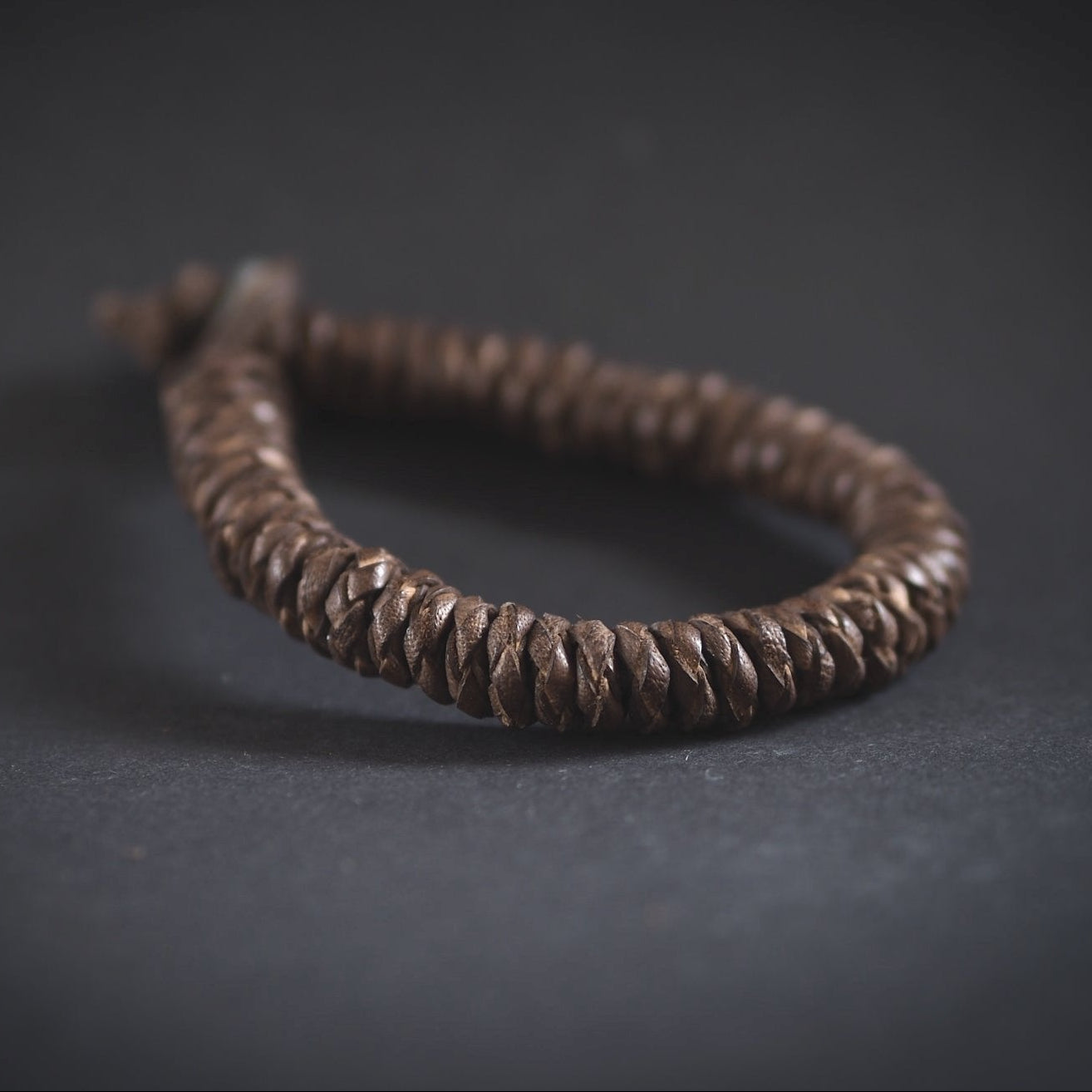 Produktfoto av brunt brokk-armband fotograferat mot mörkgrå bakgrund. Visar väl den täta flätningen och metallknappslutningen.