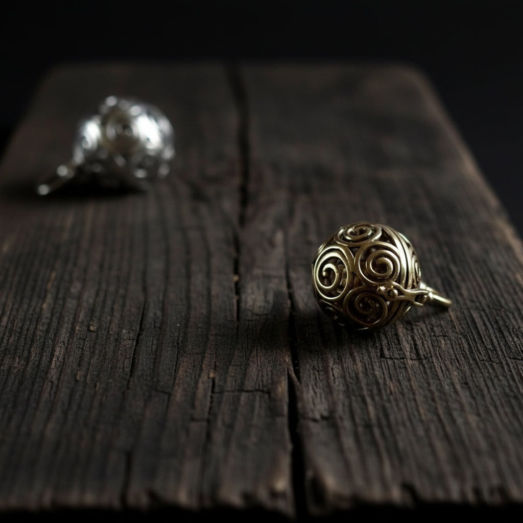 Två berlocker med solspiralmönster - en i silverpläterad finish och en i mässing. Båda visar samma intrika spiraldesign utskärd genom den sfäriska bärlocken, fotograferad från sidan för att visa mönstrets komplexitet.